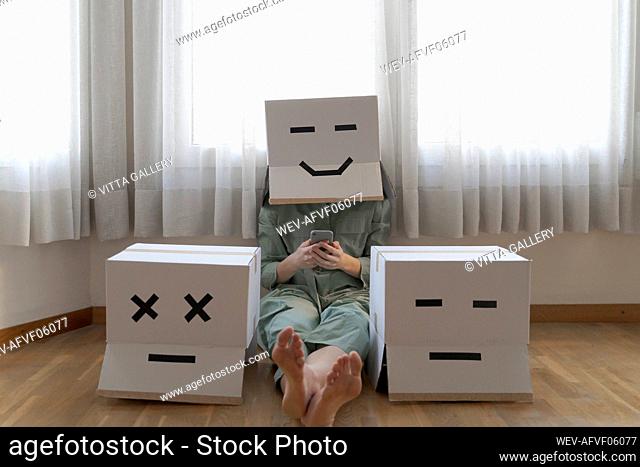 Woman wearing cardbox with smiling smiley and sitting between different cardboxes at home and using smartphone, smiling and bored cardbox
