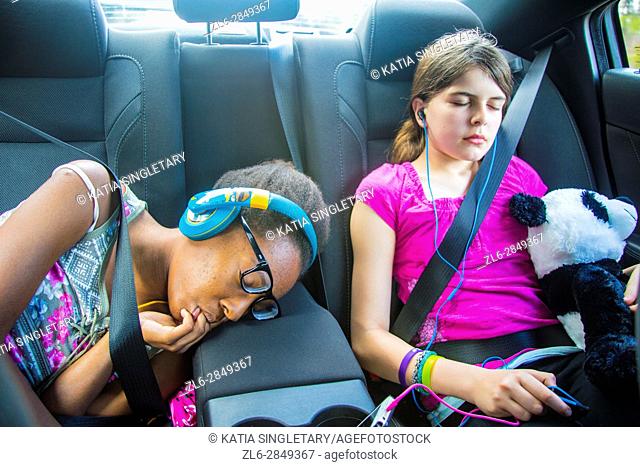 2 girls, one caucasian preteen and one african american teen sucking her fingers in the car sleeping and resting with stuff animals and earphones on