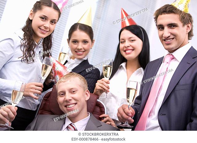 Portrait of joyful male wearing birthday cap looking at camera on background of cheerful colleagues
