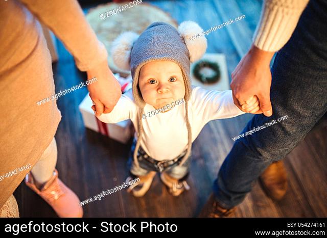 Funny little baby boy 1 year old learning walk home in winter in a decorated New Year house. Young family dad and mom hold by the hands of his son in the loft...