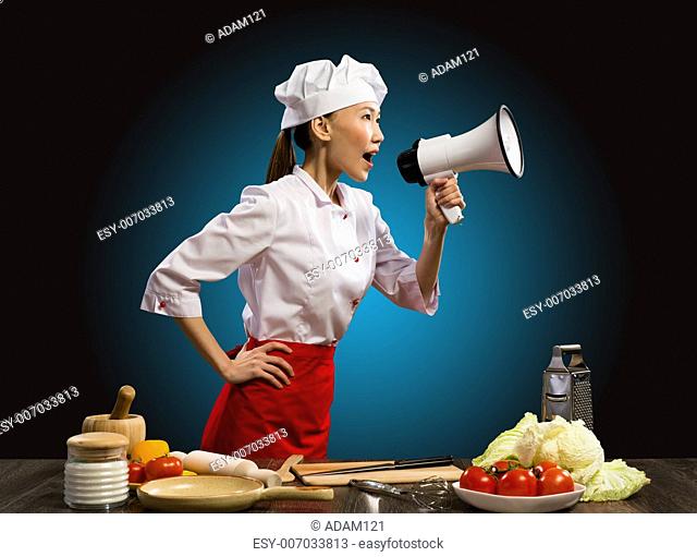 Asian female chef shouting into a megaphone, in front of her kitchen table with vegetables and items