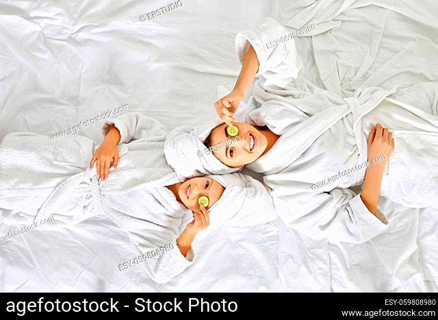 happy mom and daughter in white bathrobe lie on the bed and applying pieces of cucumber to their eyes, Top view