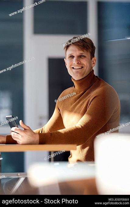 Happy businessman holding mobile phone sitting at desk in office