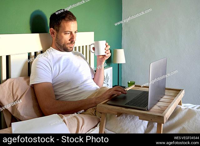 Mid adult man with coffee cup using laptop while resting in bedroom
