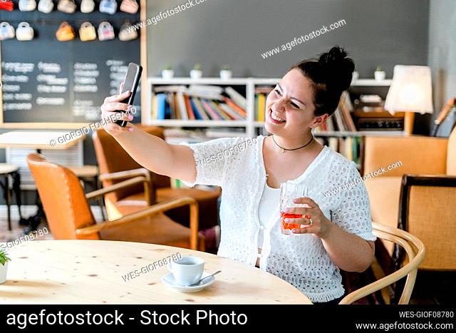 Smiling woman holding drink taking selfie with smart phone at table in cafe