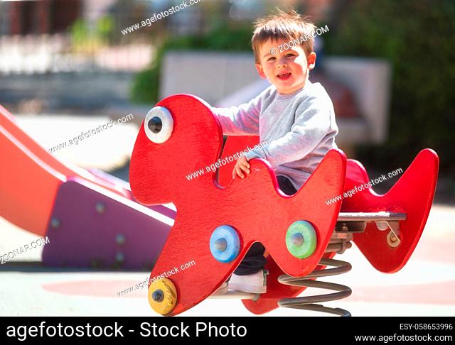 Cute happy baby boy with blond hair riding red spring rider or rocker on sunny summer day