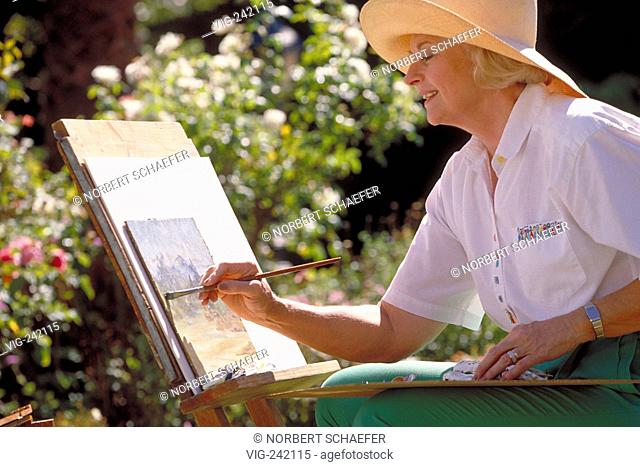 portrait, close-up, woman mid of 60 wearing green trousers, a white shirt and a strawhat sits with an easel in the garden painting  - GERMANY, 23/08/2004
