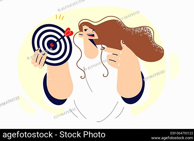 Woman holds target for darts and smiles demonstrating accurate hit in center of target. Purposeful girl dressed in casual shirt recommends striving for success...