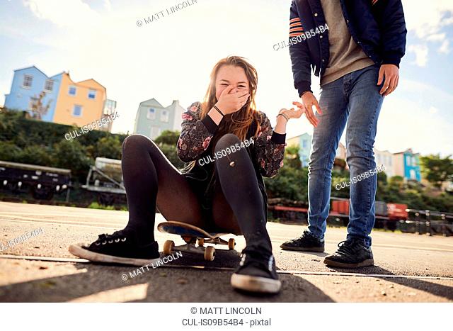 Two friends fooling around outdoors, young woman sitting on skateboard, laughing, low section, Bristol, UK