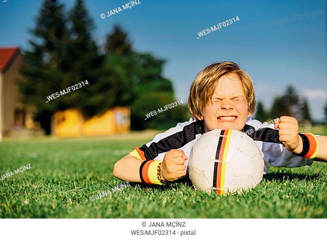 Boy in German soccer shirt lying on grass, keeping fingers crossed for world championship