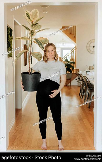 Pregnant woman holding potted plant while standing in living room at home