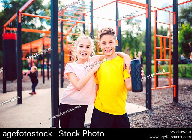 Caucasian children brother and sister stand in embrace at outdoor sports ground with smile and flask with water after training