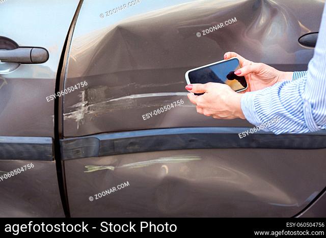Unknown woman with mobile phone in hands taking picture of damaged part of car, photographing scratches and dents on auto door