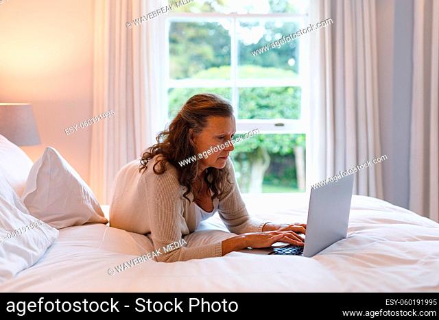 Happy senior caucasian woman in bedroom lying on bed, using laptop
