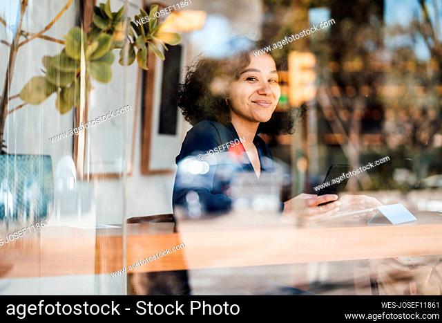 Smiling woman with smart phone seen through glass of cafe