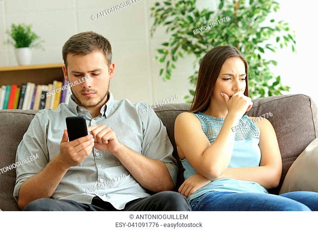 Husband addicted to smart phone watching content beside his worried wife looking down sitting on a sofa at home