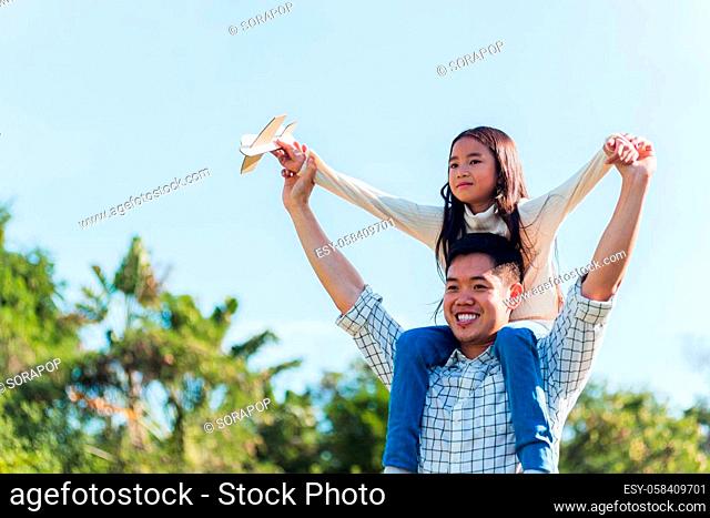 Happy Asian young family father and carrying an excited girl on shoulders having fun and enjoying outdoor lifestyle together playing aircraft toy on sunny...