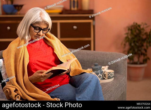 Old woman reading a book while sitting on sofa in living room