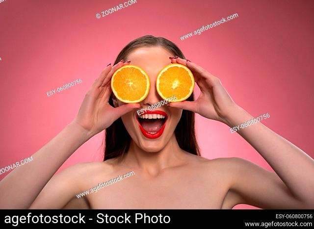 Funny beautiful beauty model young woman holding two juicy orange slices as her eyes or glasses. Charming joyful funny lady with red lips and long hair isolated...