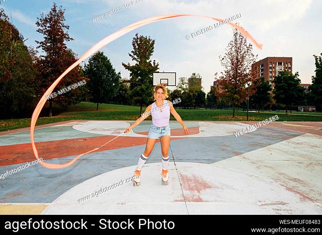 Smiling woman dancing with ribbon and roller skating at sports court