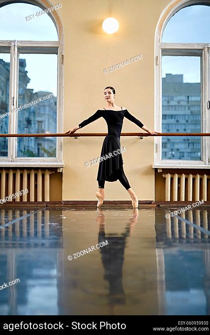 Beautiful ballerina with closed eyes stands on pointes and holds her hands on the ballet barre in the dance hall. She wears a black leotard with a skirt and...