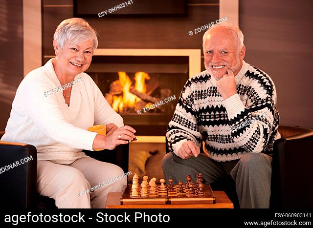 Portrait of elderly couple playing chess at home by fireplace in winter, looking at camera, smiling