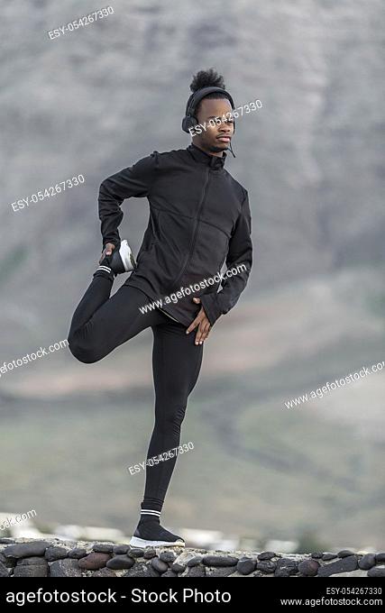 Handsome African American male in sportswear listening to music and doing warm up exercise for legs while standing on stones on blurred background of nature