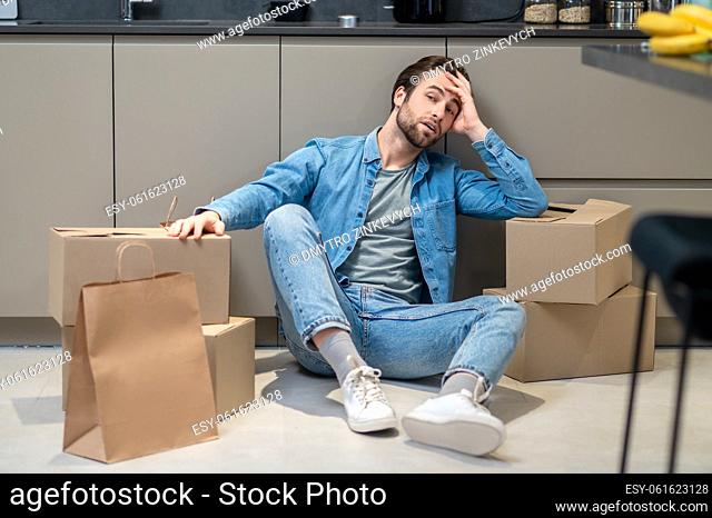 Reflections. Thoughtful man touching head with hand looking away sitting between cardboard boxes on floor in kitchen at home