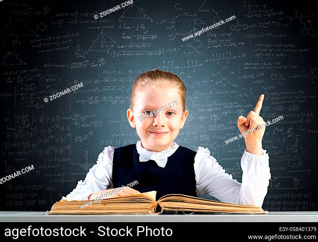 Smiling little girl sitting at desk with open book. Research and education in school. Happy schoolgirl in school wear finger pointing up