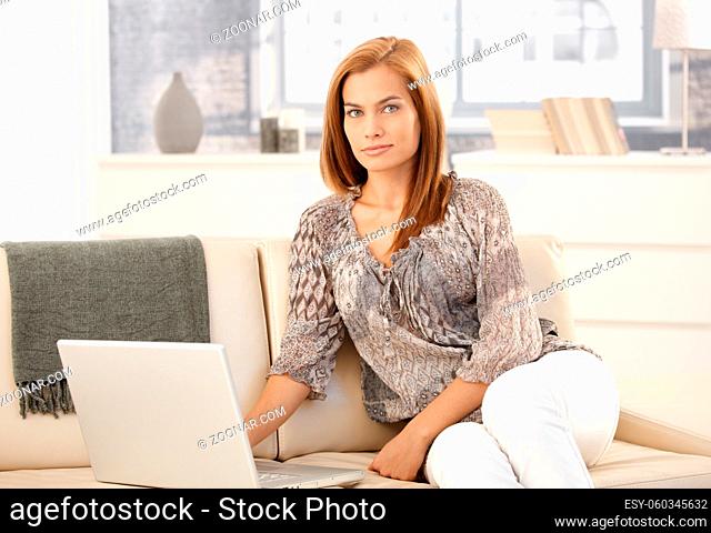 Portrait of attractive woman sitting on couch with laptop computer, looking at camera, smiling