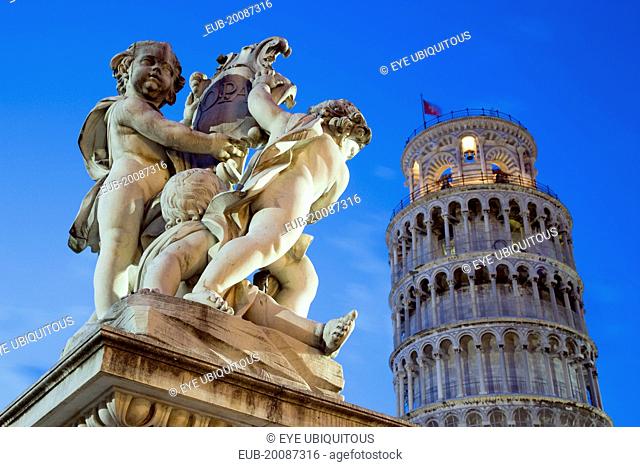 An illuminated statue in The Piazza del Duomo of cherubs holding a shield bearing the Cross of Pisa with the Leaning Tower beyond all within the UNESCO World...
