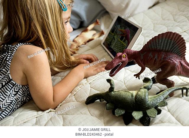 Caucasian girl laying on bed with toy dinosaurs using digital tablet