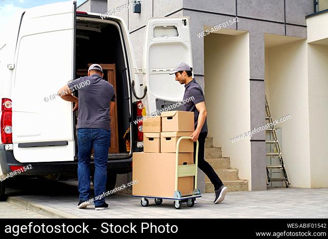 Man opening door while colleague pushing cart with boxes near van