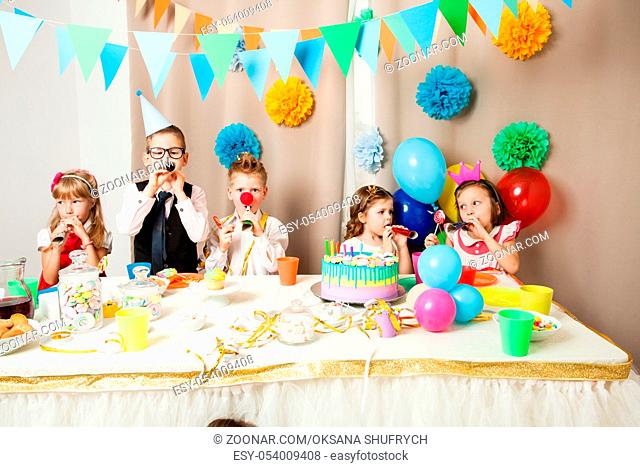 Group of smiling children playing on the birthday party in decorated room. Happy kids blowing in pipes on birthday party