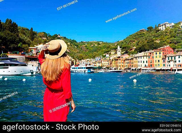 Tourism in Italy. Back view of beautiful young woman in red dress looks towards Portofino colorful village with yachts moored in its harbor, Portofino, Liguria