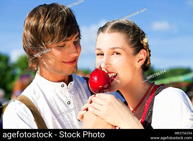 Young and beautiful couple in traditional Bavarian Tracht - Dirndl and Lederhosen - embracing each other on a fair like a Dult or the Oktoberfest eating...