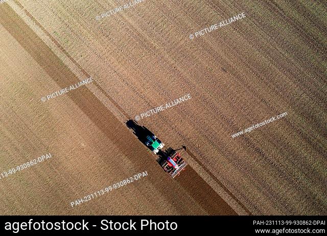 13 November 2023, Mecklenburg-Western Pomerania, Wessin: A tractor pulls a seed drill across a field and sows seed. (Aerial shot with a drone) Farmers in...