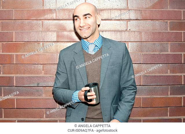 Portrait of young businessman drinking coffee whilst leaning against brick wall