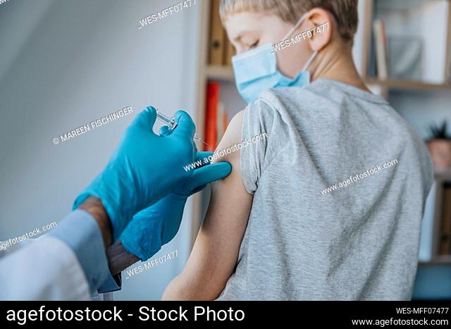 Boy taking COVID-19 vaccine on arm while sitting by doctor at clinic