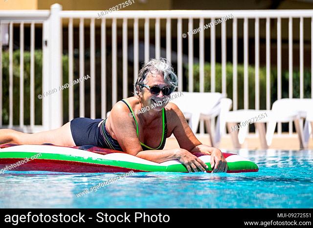 Happy cheerful caucasian people old senior woman lay down on coloured trendy lilo enjoying the swimmin pool outdoor leisure activity in summer time for vacation...