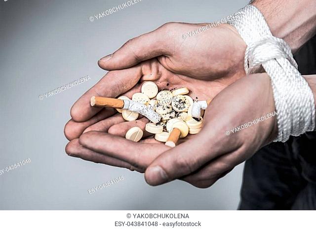Top view close up of arms of young man keeping pills and cigarette butts in palms. He is tied by rope. Drug and smoking addiction concept