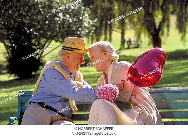 park-scene, outdoor, senior-couple wearing bright dresses and strawhats sits with a heart shaped balloon on a parkbench flirting  - GERMANY, 23/08/2004