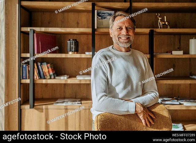 Happy mature man standing at chair against shelf in living room