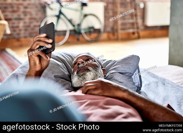 Man with white beard using smart phone lying on bed at home