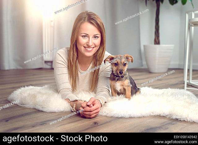 Attractive friendly woman posing with her small dog on a fluffy rug on a wood floor in the living room smiling happily at the camera
