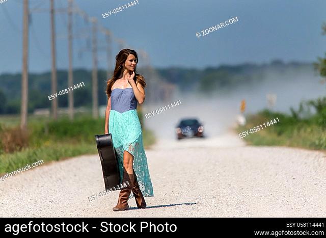 Female walking down a dirt road hitchhiking with a guitar case