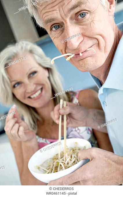 Close-up of a senior man eating spaghetti with a senior woman smiling in the background