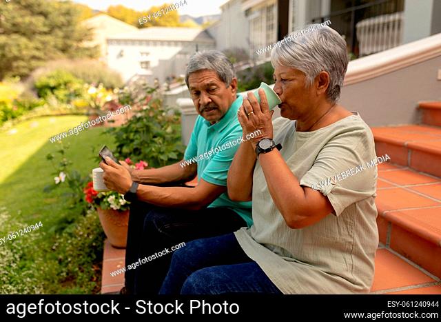 Biracial senior woman drinking coffee and senior man using digital tablet while sitting on steps