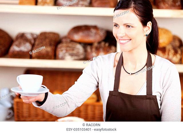 Friendly young woman in an apron serving a hot cup of coffee in a bakery against a backdrop of specialist loaves of bread