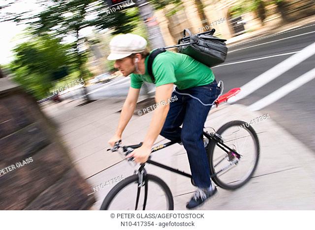 Young  man, wearing a cap, an earphone, and balancing his shoulder bag on his back, riding a bicycle on a city sidewalk